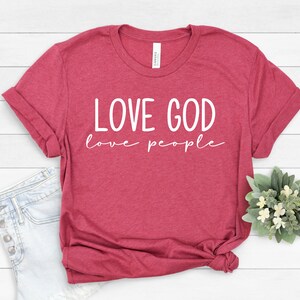 K&ouml;nnte beinhalten: Ein weinrotes T-Shirt mit dem wei&szlig;en Aufdruck "LOVE GOD love people". Das Shirt ist gefaltet und hat hochgekrempelte &Auml;rmel. Dazu geh&ouml;ren hellblaue Jeansshorts und ein kleines Blumenarrangement.