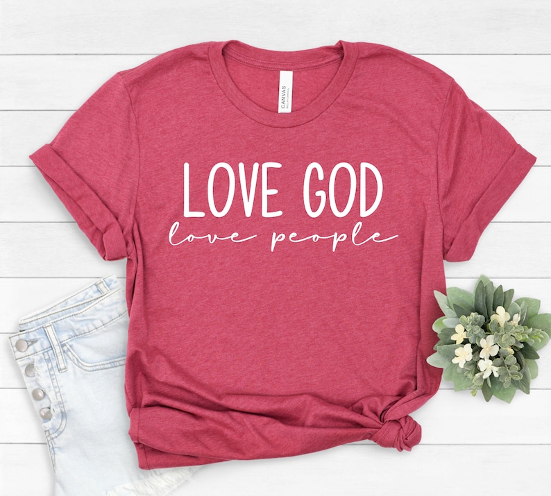 K&ouml;nnte beinhalten: Ein weinrotes T-Shirt mit dem wei&szlig;en Aufdruck "LOVE GOD love people". Das Shirt ist gefaltet und hat hochgekrempelte &Auml;rmel. Dazu geh&ouml;ren hellblaue Jeansshorts und ein kleines Blumenarrangement.