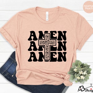 May include: A heather peach t-shirt with a black graphic of the word "Amen" repeated three times with a cross in the center. The cross is decorated with a leopard print pattern.