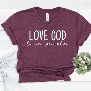 K&ouml;nnte beinhalten: Ein weinrotes T-Shirt mit hochgekrempelten &Auml;rmeln. Das Shirt zeigt den wei&szlig;en Text "LOVE GOD love people" in Gro&szlig;buchstaben und Kursivschrift. Das Shirt wird mit Jeansshorts und einem kleinen Blumenarrangement pr&auml;sentiert.