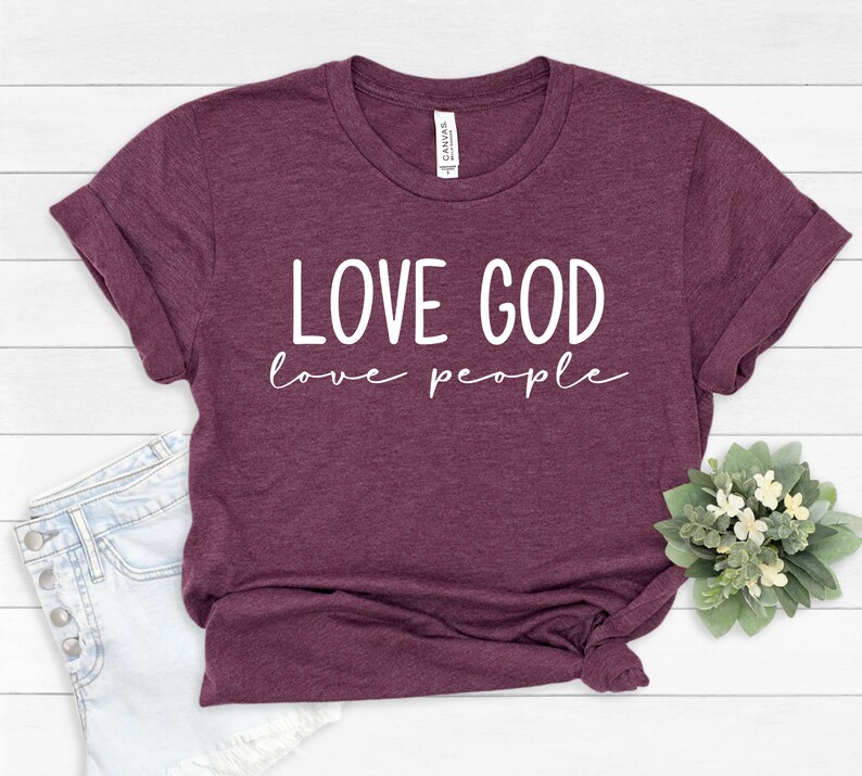 K&ouml;nnte beinhalten: Ein weinrotes T-Shirt mit hochgekrempelten &Auml;rmeln. Das Shirt zeigt den wei&szlig;en Text "LOVE GOD love people" in Gro&szlig;buchstaben und Kursivschrift. Das Shirt wird mit Jeansshorts und einem kleinen Blumenarrangement pr&auml;sentiert.