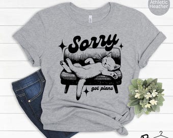 May include: A gray t-shirt with a black and white illustration of a cat sleeping on a couch. The text "Sorry got plans" is printed on the shirt.
