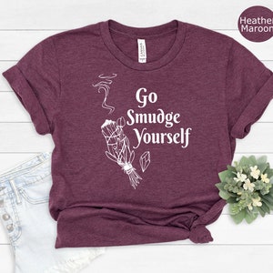 K&ouml;nnte beinhalten: Ein T-Shirt in Heide-Maroon mit dem Text "Go Smudge Yourself" in Wei&szlig;. Das Shirt zeigt eine wei&szlig;e Illustration eines R&auml;ucherstabs mit Rauch, der dar&uuml;ber aufsteigt.
