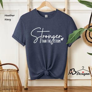 Może przedstawiać: Koszulka w kolorze granatowym z domieszką hebanu z napisem "Stronger than the storm" w białym kolorze z małym symbolem serca.