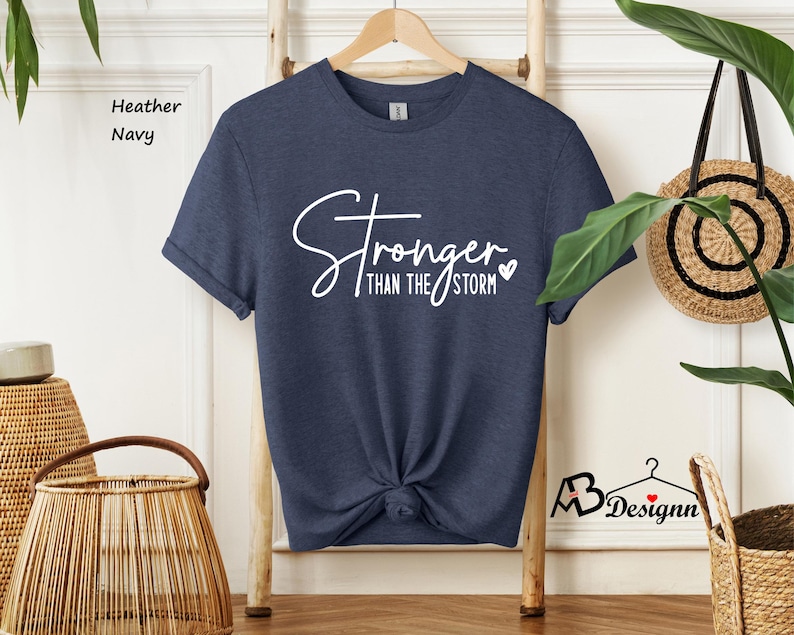 Może przedstawiać: Koszulka w kolorze granatowym z domieszką hebanu z napisem "Stronger than the storm" w białym kolorze z małym symbolem serca.