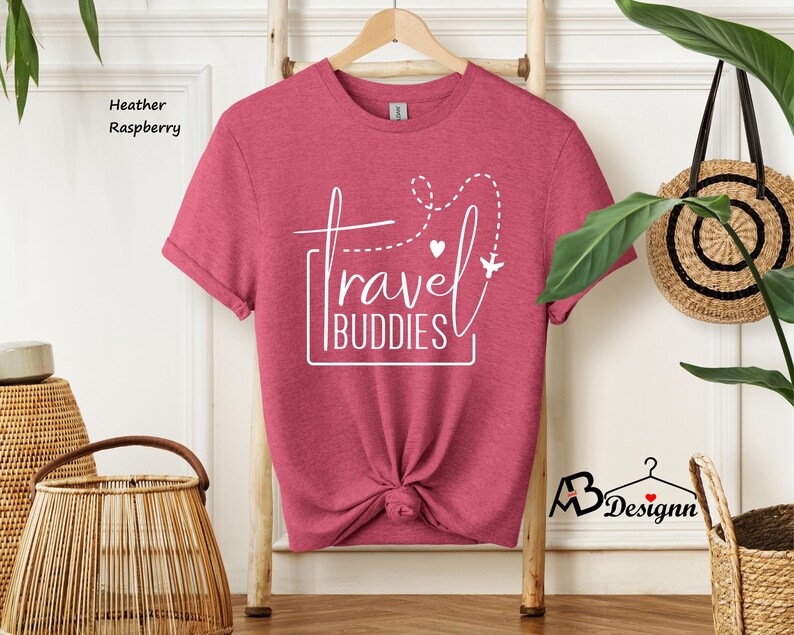 K&ouml;nnte beinhalten: Ein kurz&auml;rmeliges T-Shirt in Himbeer-Melange mit einem wei&szlig;en Grafikdruck, der "travel BUDDIES" zeigt, erg&auml;nzt durch eine gestrichelte Flugroute, ein kleines Herz und ein Flugzeugsymbol. Das T-Shirt h&auml;ngt auf einem Holzkleiderb&uuml;gel.