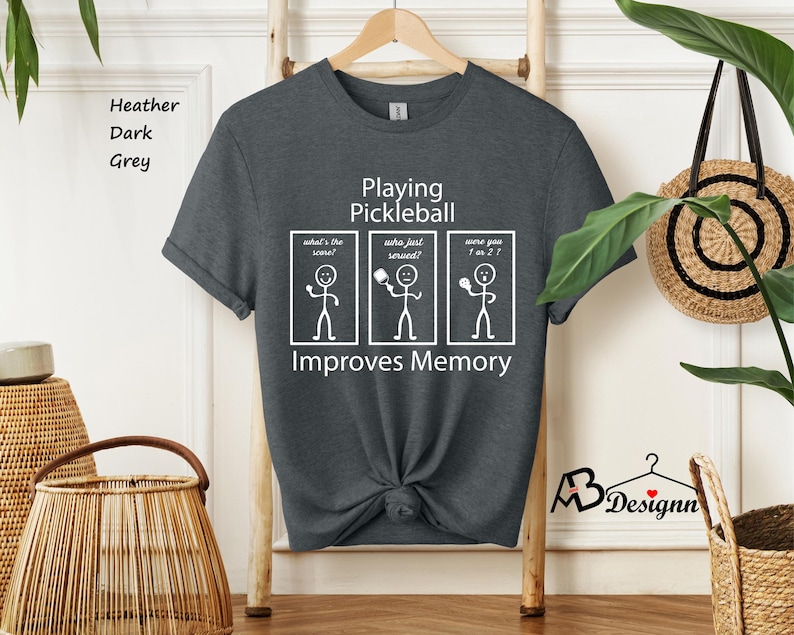 May include: Dark heather grey t-shirt with a white graphic that says "Playing Pickleball Improves Memory". The graphic features three stick figures with different pickleball-related questions.