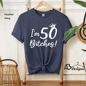 May include: A heather navy short-sleeve t-shirt featuring white screen-printed text that reads I'm 50 Bitches! with a small crown graphic above the number 50. The shirt has a crew neckline and is displayed on a wooden hanger against a light background.