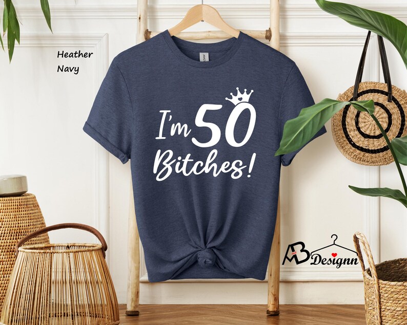 May include: A heather navy short-sleeve t-shirt featuring white screen-printed text that reads I'm 50 Bitches! with a small crown graphic above the number 50. The shirt has a crew neckline and is displayed on a wooden hanger against a light background.