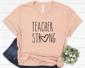 May include: A peach-colored t-shirt with the text "TEACHER STRONG" in black lettering with a heart in the middle of the word "STRONG".