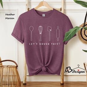 May include: A heather maroon t-shirt with a white graphic of kitchen utensils and the text "Let's Dough This!"