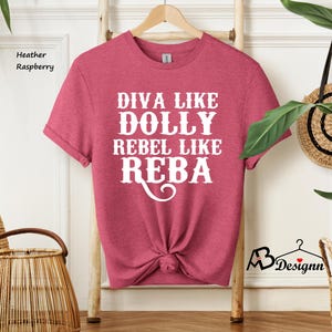 Puede incluir: Una camiseta de manga corta color frambuesa jaspeado con el texto en blanco DIVA LIKE DOLLY REBEL LIKE REBA. La camiseta presenta un cuello redondo y se muestra en una percha de madera. La tela tiene una textura jaspeada suave en toda la prenda.
