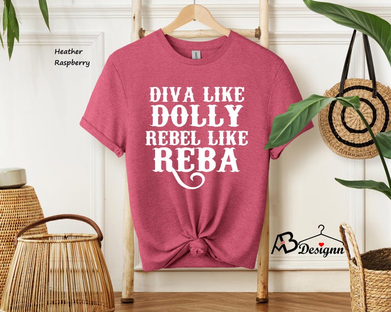 Puede incluir: Una camiseta de manga corta color frambuesa jaspeado con el texto en blanco DIVA LIKE DOLLY REBEL LIKE REBA. La camiseta presenta un cuello redondo y se muestra en una percha de madera. La tela tiene una textura jaspeada suave en toda la prenda.