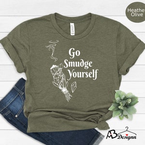 K&ouml;nnte beinhalten: Ein gr&uuml;nes T-Shirt mit dem Text "Go Smudge Yourself" in Wei&szlig;. Das Shirt zeigt eine wei&szlig;e Illustration eines R&auml;ucherstabs mit aufsteigendem Rauch.