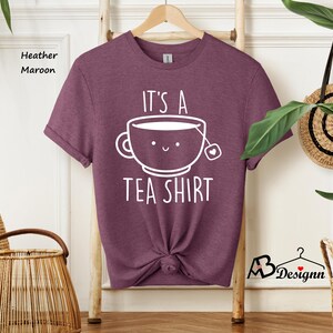 May include: Heather maroon t-shirt with the text "It's a Tea Shirt" and a smiling teacup graphic. The shirt is displayed on a wooden hanger. The shirt is knotted at the waist.