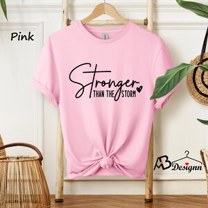 Może przedstawiać: R&oacute;żowa koszulka z napisem "Stronger than the storm" czarnymi literami z małym sercem.