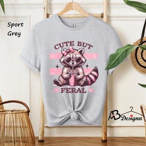 Op de afbeelding: Een lichtgrijs T-shirt met een schattig cartoon-wasbeer die een zonnebril draagt en een roze drankje drinkt. De tekst "Cute But Feral" staat op het shirt gedrukt.