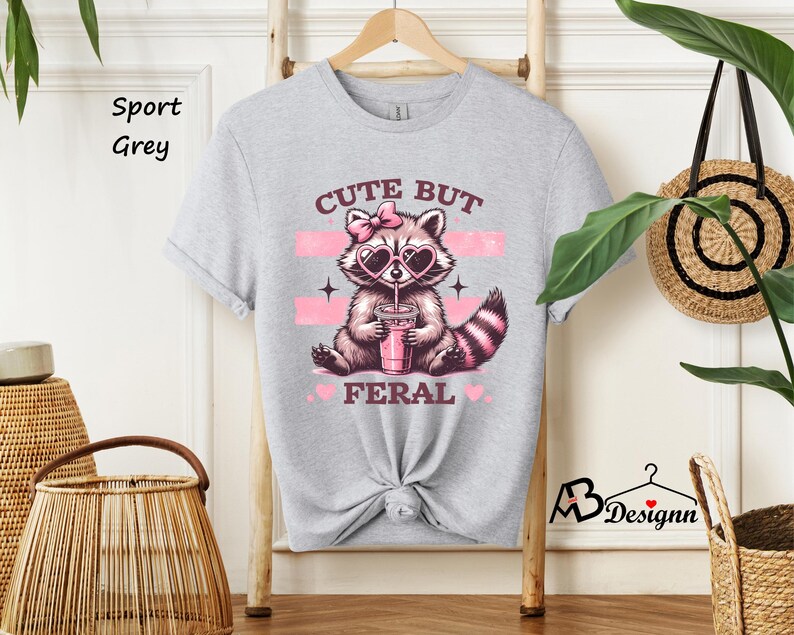 Op de afbeelding: Een lichtgrijs T-shirt met een schattig cartoon-wasbeer die een zonnebril draagt en een roze drankje drinkt. De tekst "Cute But Feral" staat op het shirt gedrukt.