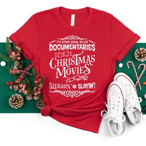 May include: A red t-shirt with white text that reads "It's Either Serial Killer Documentaries or Christmas Movies Sleighin' or Slayin'!" The shirt is styled with Christmas decorations and white sneakers.