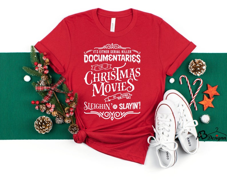 May include: A red t-shirt with white text that reads "It's Either Serial Killer Documentaries or Christmas Movies Sleighin' or Slayin'!" The shirt is styled with Christmas decorations and white sneakers.