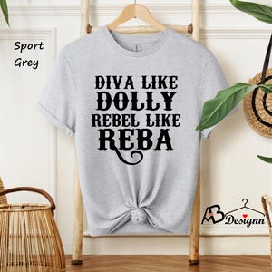 Puede incluir: Una camiseta de manga corta en color gris sport con texto negro que dice DIVA LIKE DOLLY REBEL LIKE REBA. La prenda tiene cuello redondo y se muestra en una percha de madera. El dise&ntilde;o est&aacute; centrado en el pecho con una fuente decorativa.