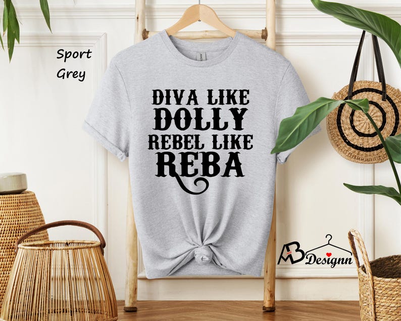 Puede incluir: Una camiseta de manga corta en color gris sport con texto negro que dice DIVA LIKE DOLLY REBEL LIKE REBA. La prenda tiene cuello redondo y se muestra en una percha de madera. El dise&ntilde;o est&aacute; centrado en el pecho con una fuente decorativa.
