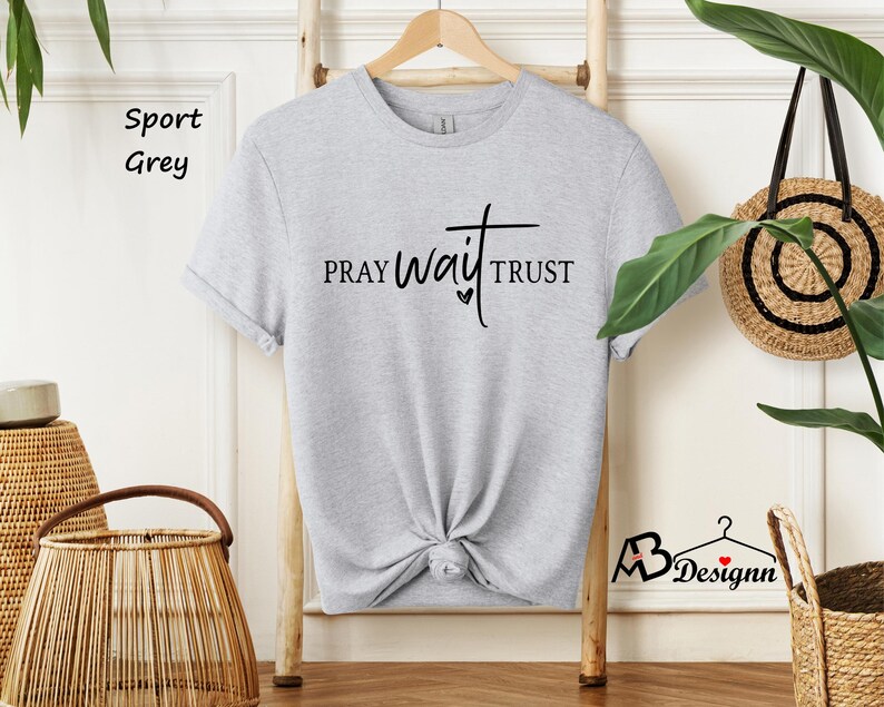 May include: A light gray t-shirt with the words "PRAY wait TRUST" in black script. The shirt is hanging on a wooden hanger against a white wall. The words "Sport Grey" are printed in black in the upper left corner.