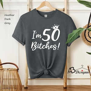 May include: A heather dark grey short-sleeve t-shirt featuring white screen-printed text that reads I'm 50 Bitches! with a small crown icon above the number 50. The shirt has a crew neckline and is displayed on a wooden hanger for presentation.