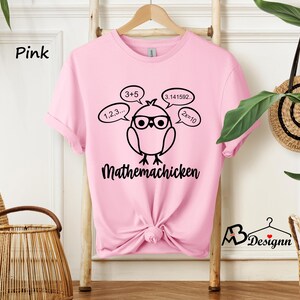 Op de afbeelding: Roze T-shirt met een zwarte afbeelding van een cartoonkip met een bril en gedachtebellen met wiskundige vergelijkingen. De tekst "Mathemachicken" staat onder de kip.