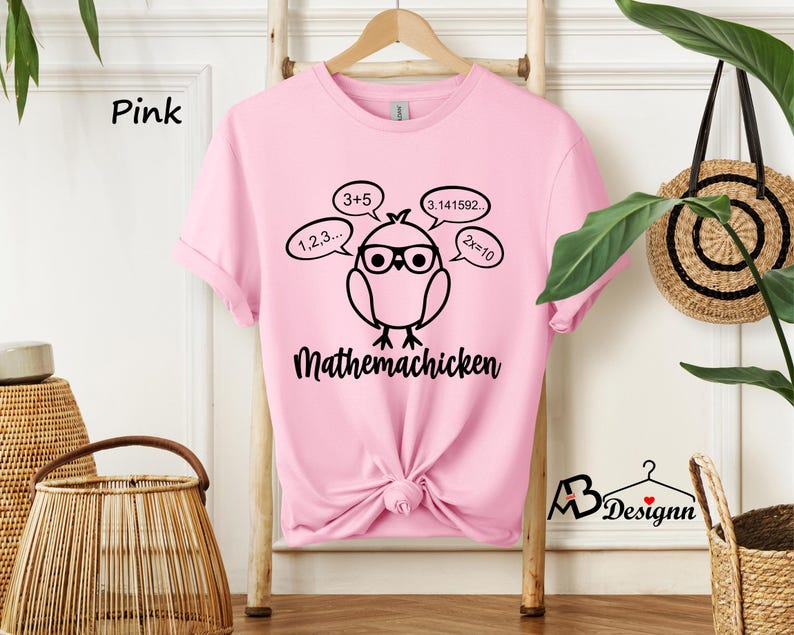Op de afbeelding: Roze T-shirt met een zwarte afbeelding van een cartoonkip met een bril en gedachtebellen met wiskundige vergelijkingen. De tekst "Mathemachicken" staat onder de kip.