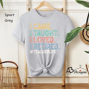 Może przedstawiać: Jasnoszara koszulka z napisem "I CAME. I TAUGHT. I LOVED. I RETIRED. #TEACHERLIFE" w kolorowych literach. Koszulka wisi na drewnianym wieszaku. Koszulka jest zawiązana na dole.