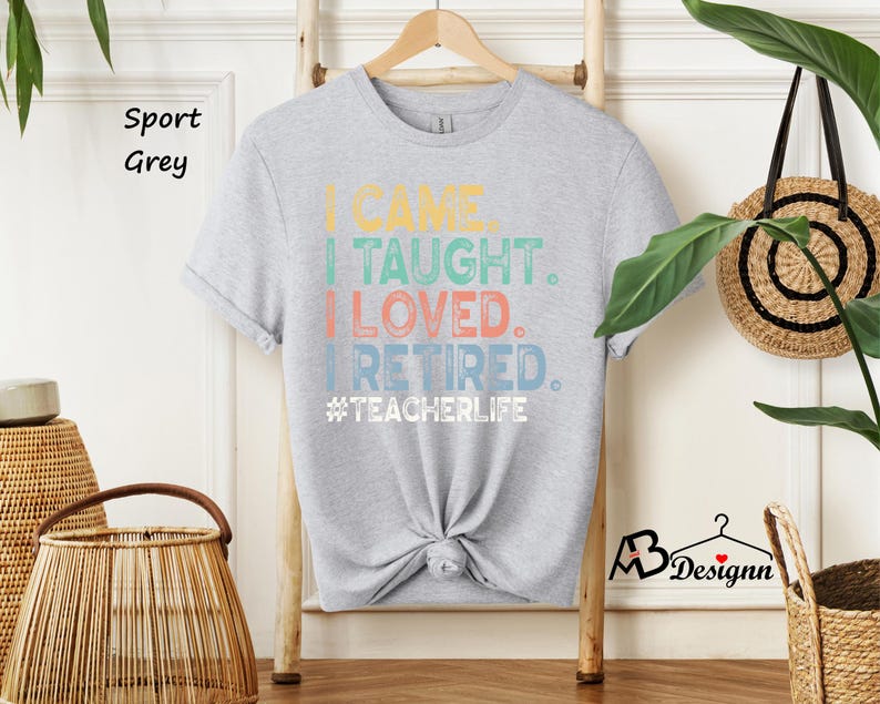 Może przedstawiać: Jasnoszara koszulka z napisem "I CAME. I TAUGHT. I LOVED. I RETIRED. #TEACHERLIFE" w kolorowych literach. Koszulka wisi na drewnianym wieszaku. Koszulka jest zawiązana na dole.