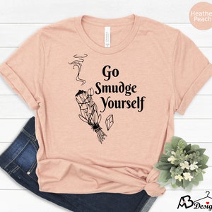 K&ouml;nnte beinhalten: Ein T-Shirt in der Farbe Heidelbeer-Pfirsich mit einem schwarzen Aufdruck eines R&auml;ucherstabs und dem Text "Go Smudge Yourself".