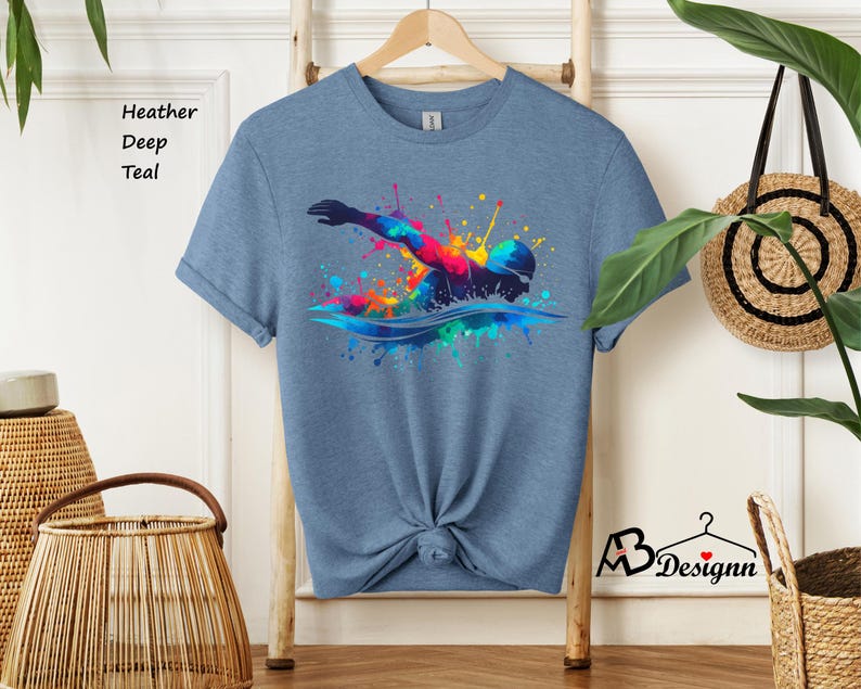 May include: A heather deep teal t-shirt with a colorful graphic of a swimmer in a splash of water. The graphic is made up of various colors, including red, orange, yellow, green, blue, and purple.