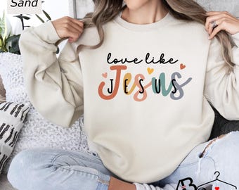 May include: Sand-colored sweatshirt with the text "love like Jesus" in black script above the word "Jesus" in colorful, rounded letters. Small heart graphics are scattered around the text. The sweatshirt has long sleeves.