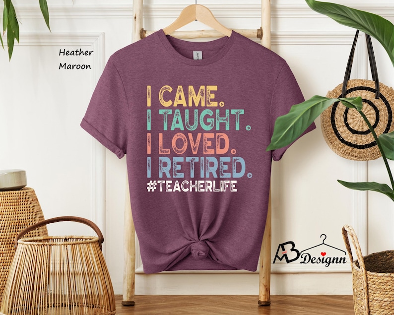 Może przedstawiać: Koszulka w kolorze wrzosowym, bordowym z napisem "I CAME. I TAUGHT. I LOVED. I RETIRED. #TEACHERLIFE" w kolorze ż&oacute;łtym, turkusowym, koralowym i niebieskim. Koszulka jest zawiązana na dole. Drewniana drabina i wiklinowy kosz w tle.