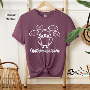 Op de afbeelding: Een heather maroon t-shirt met een witte afbeelding van een cartoonkip met een bril en gedachtebellen met wiskundige vergelijkingen. De tekst "Mathemachicken" staat onder de kip.