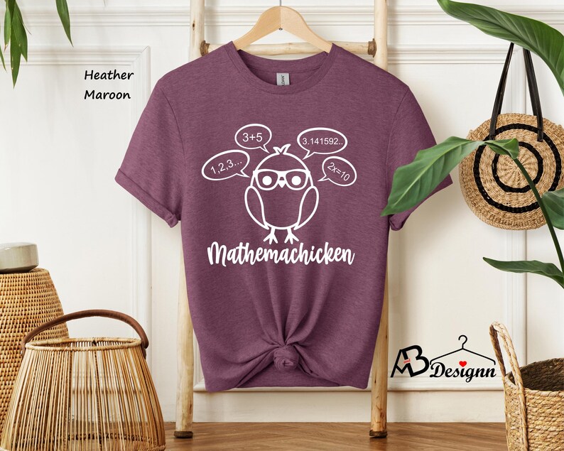 Op de afbeelding: Een heather maroon t-shirt met een witte afbeelding van een cartoonkip met een bril en gedachtebellen met wiskundige vergelijkingen. De tekst "Mathemachicken" staat onder de kip.