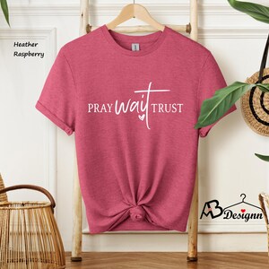 May include: Heather raspberry t-shirt with the words "PRAY WAIT TRUST" in white script. The shirt is knotted at the bottom and hangs on a wooden hanger. The words are in a stylized font with a heart symbol.
