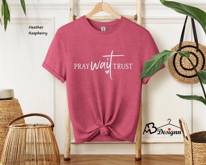 May include: Heather raspberry t-shirt with the words "PRAY WAIT TRUST" in white script. The shirt is knotted at the bottom and hangs on a wooden hanger. The words are in a stylized font with a heart symbol.