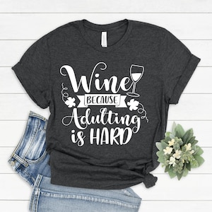 Pu&ograve; includere: T-shirt grigio scuro con la scritta bianca "Wine Because Adulting is HARD". Il design include un calice di vino e decorazioni. La maglietta &egrave; piegata, con un paio di jeans blu visibili.
