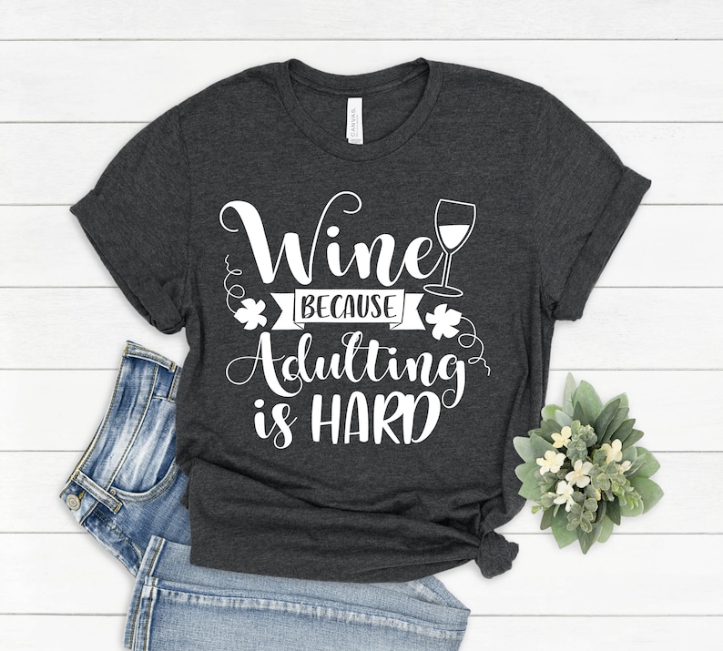Pu&ograve; includere: T-shirt grigio scuro con la scritta bianca "Wine Because Adulting is HARD". Il design include un calice di vino e decorazioni. La maglietta &egrave; piegata, con un paio di jeans blu visibili.