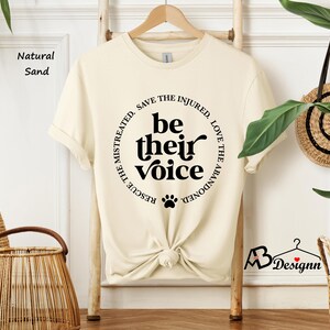 May include: A cream-colored t-shirt with the text "be their voice" in a black circular design. The design includes a paw print and the phrases "Rescue the mistreated, save the injured, love the abandoned". The shirt is knotted at the waist.