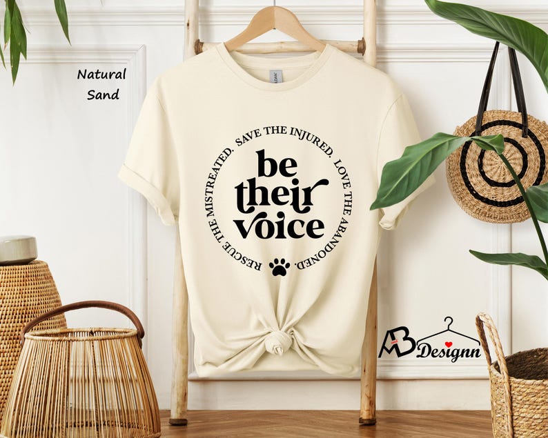 May include: A cream-colored t-shirt with the text "be their voice" in a black circular design. The design includes a paw print and the phrases "Rescue the mistreated, save the injured, love the abandoned". The shirt is knotted at the waist.