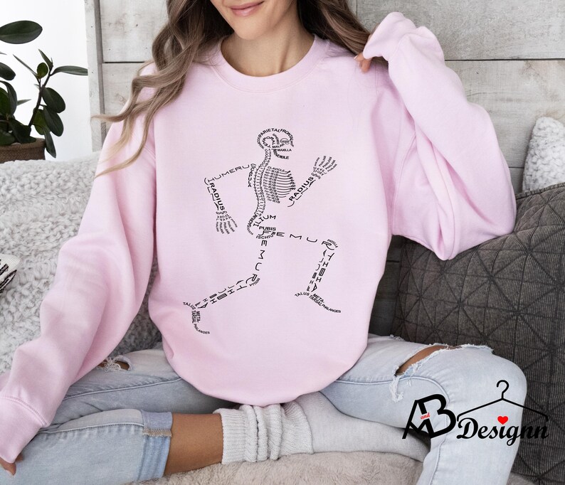 Peut inclure: Sweat-shirt rose clair avec un motif de coureur squelettique noir. Les os sont &eacute;tiquet&eacute;s avec des termes anatomiques. Le sweat-shirt a des manches longues et un col rond. Le motif est centr&eacute; sur le devant.