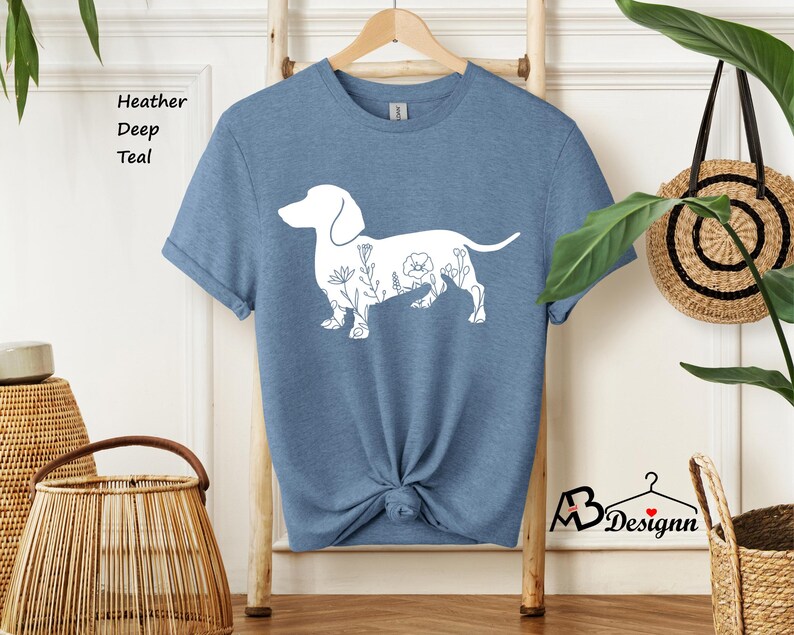 May include: A heather deep teal t-shirt featuring a white outline of a dachshund with floral details. The shirt is on a wooden hanger.