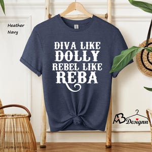 Puede incluir: Una camiseta de manga corta en color azul marino jaspeado con el texto en blanco que dice DIVA LIKE DOLLY REBEL LIKE REBA. La camiseta tiene cuello redondo y est&aacute; colgada en una percha de madera. La tela tiene una textura visible.