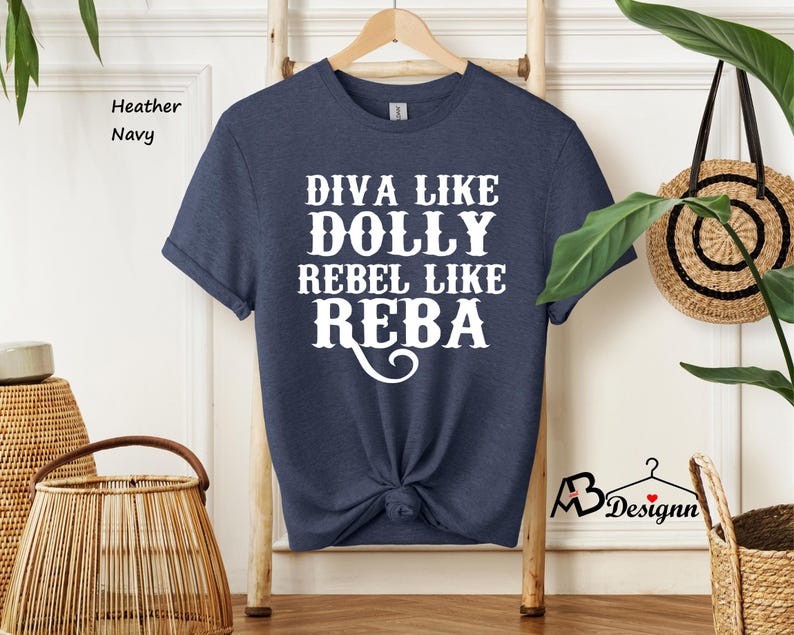 Puede incluir: Una camiseta de manga corta en color azul marino jaspeado con el texto en blanco que dice DIVA LIKE DOLLY REBEL LIKE REBA. La camiseta tiene cuello redondo y est&aacute; colgada en una percha de madera. La tela tiene una textura visible.