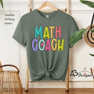 May include: Heather military green t-shirt with the words "MATH COACH" in colorful bubble letters. The shirt has a small pink heart in the letter "O". The shirt is knotted at the bottom.