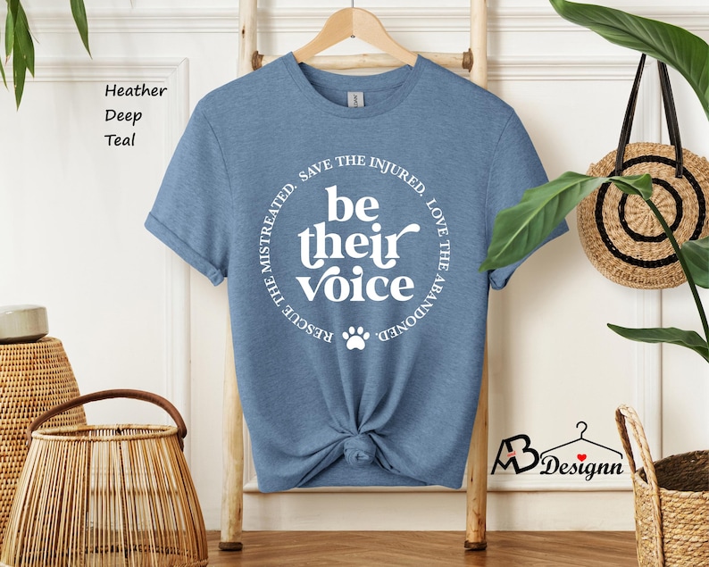 May include: A heather deep teal t-shirt with a white circular graphic that reads "be their voice" with a paw print. The text "Rescue the mistreated, save the injured, love the abandoned" surrounds the central text. The shirt is knotted at the waist.